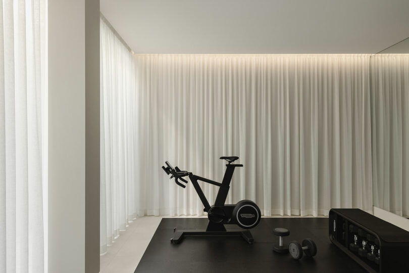 A minimal home gym with a stationary bike, two dumbbells, and a small shelf, set against floor-to-ceiling white curtains.