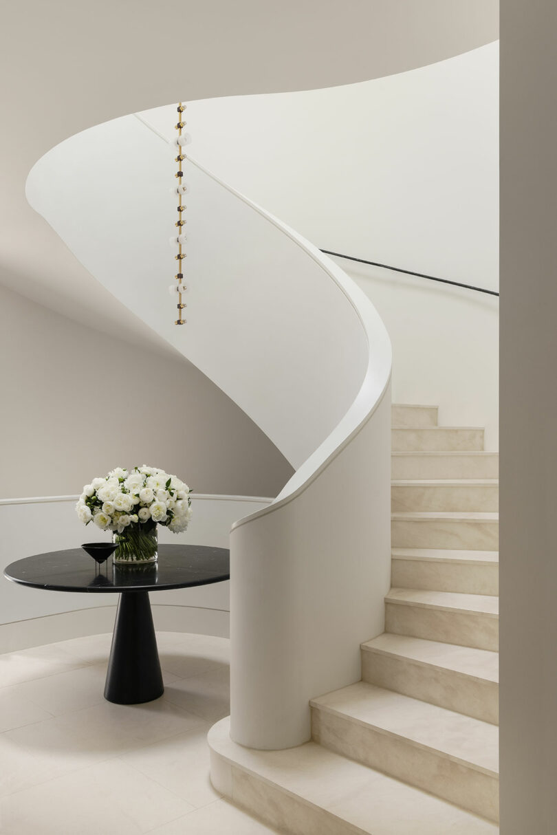 A curved white staircase with beige steps, a black round table with a white flower arrangement, and a modern hanging light fixture.