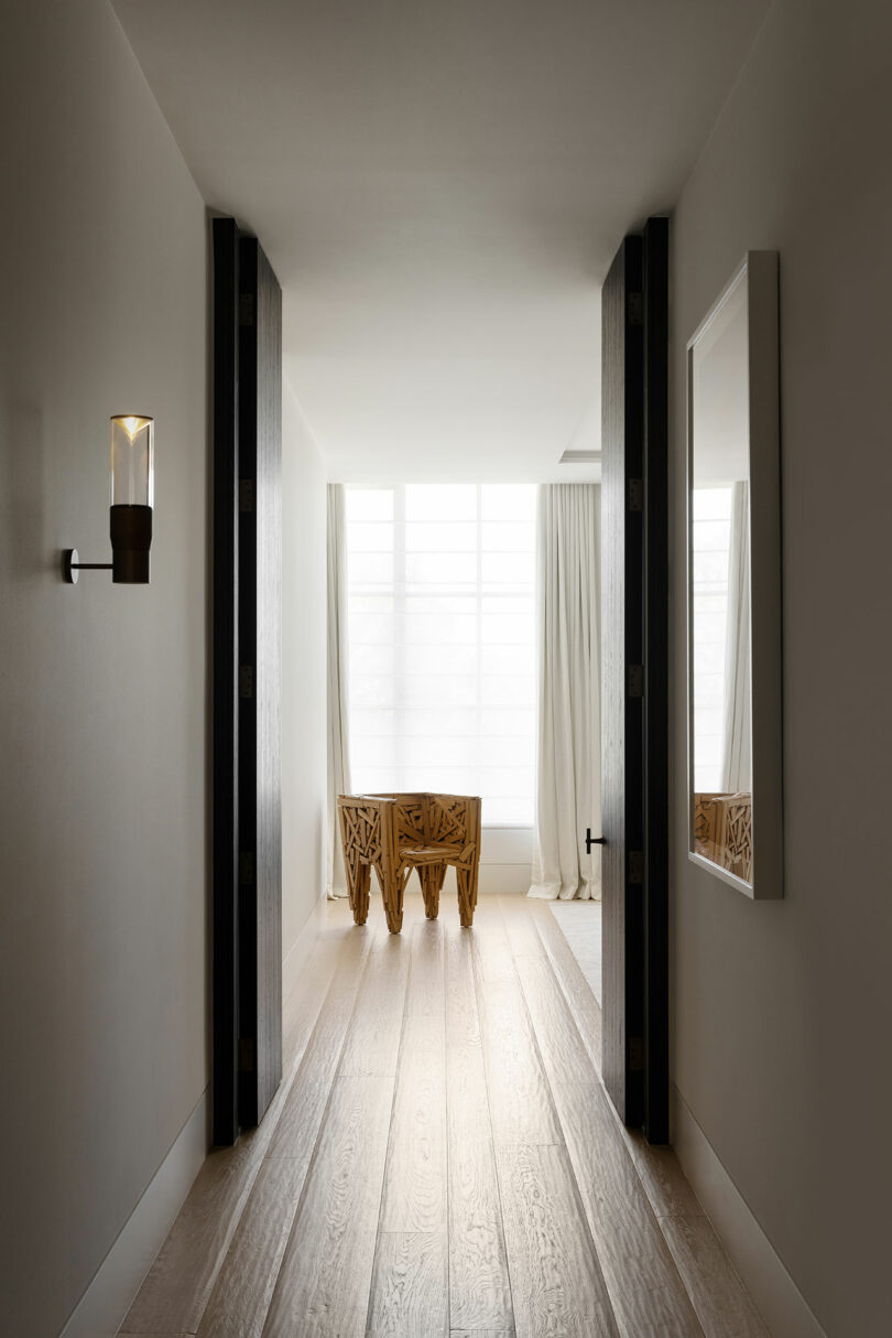 A hallway with light wood flooring leads to a bright room featuring a carved wooden table near a large window with sheer white curtains.