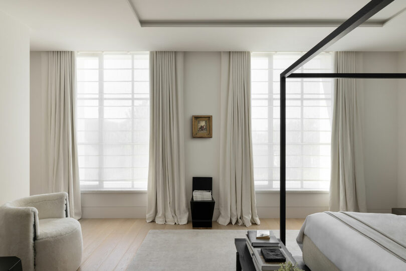 Minimalist bedroom with light-filled windows, sheer curtains, a modern black chair, neutral furnishings, and a small framed artwork between the windows.