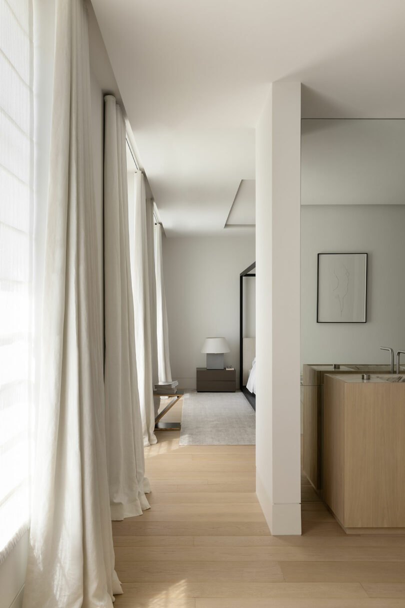 A minimal bedroom with light wood floors, tall curtains, a canopy bed, a bedside table with a lamp, and a partial view of a bathroom vanity with a sink.