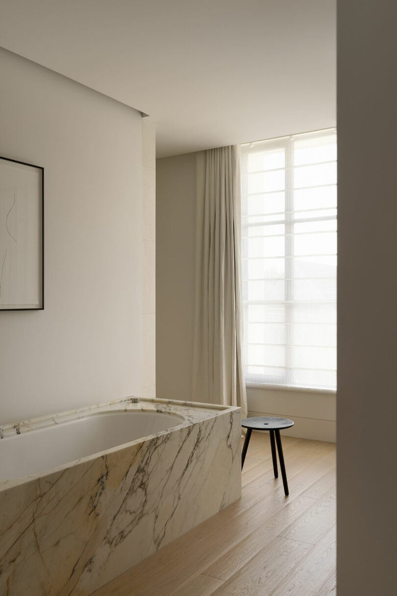 A marble bathtub sits next to a window with sheer curtains in a minimalist bathroom, featuring light wood flooring and a small black stool.