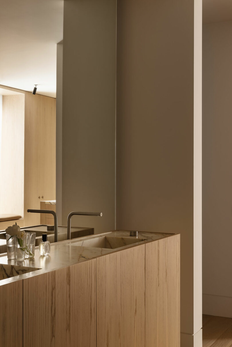 A modern bathroom with a light wood vanity, a built-in sink, a mounted faucet, a mirror, and minimal decor including a small vase with flowers and a glass.