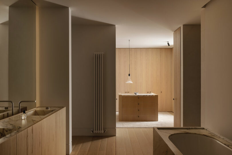 Minimalist bathroom interior with light wood cabinetry, marble countertops, a built-in bathtub, and soft ambient lighting. A pendant light hangs over a central island.