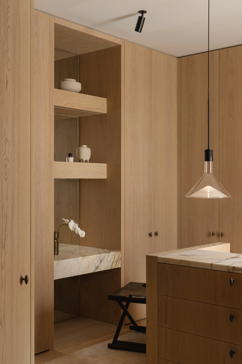 Minimalist wooden bathroom with built-in shelves, a marble countertop, a small stool, and modern pendant lighting. Decorative ceramics and a white orchid are displayed on the shelves.