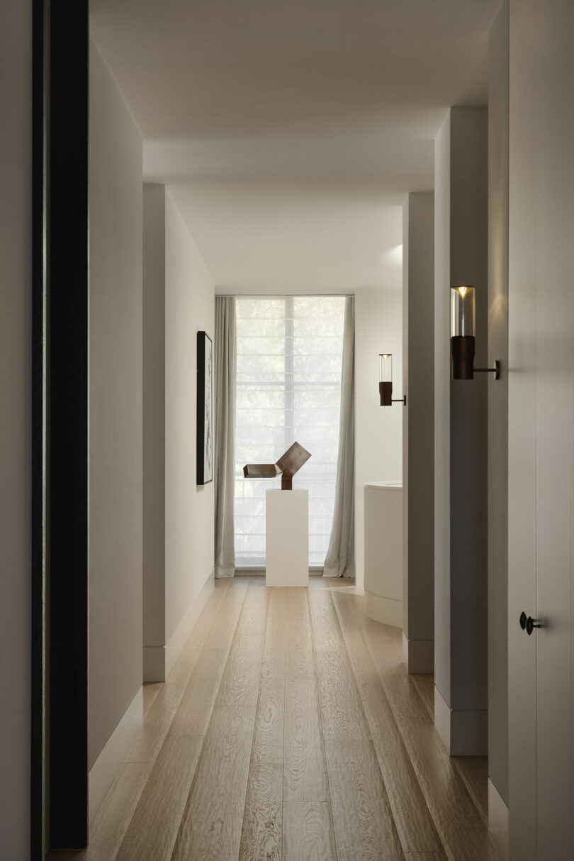 A minimalist hallway with light wood flooring, white walls, a tall window with sheer curtains, and a modern sculpture on a pedestal at the end.