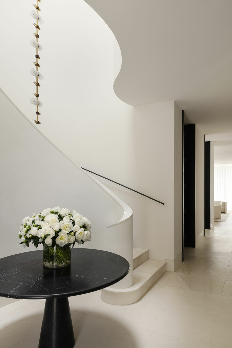 Modern interior with a curved staircase, a black round table with a vase of white flowers, and neutral-toned walls and flooring. Natural light enters from a hallway in the background.