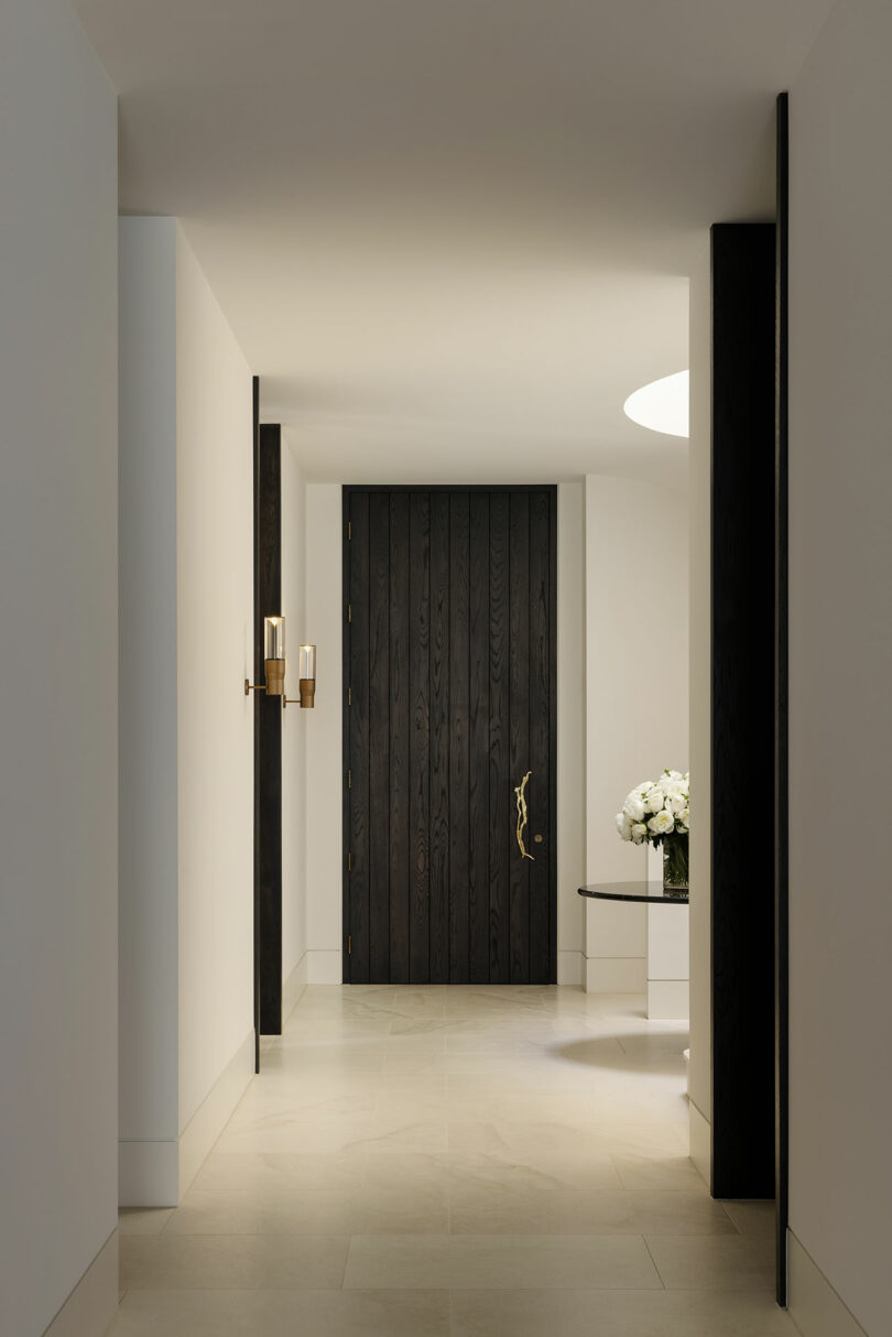 A minimalist hallway with white walls, light tile flooring, a dark wooden door, brass wall sconce, and a table with a white floral arrangement.