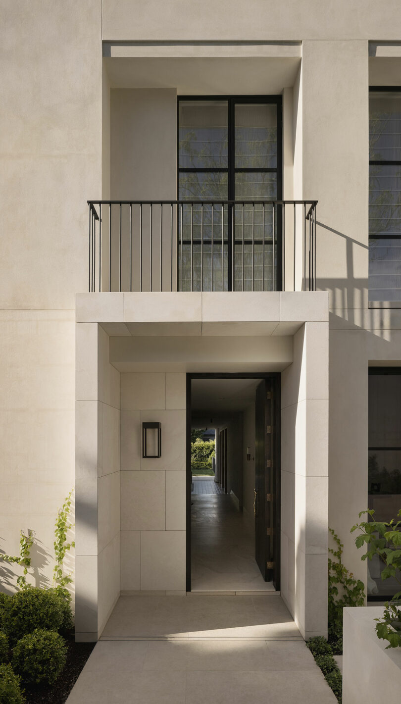 A modern beige stone building entrance with an open black door, small overhang, wall-mounted light, balcony above, and minimalist landscaping.