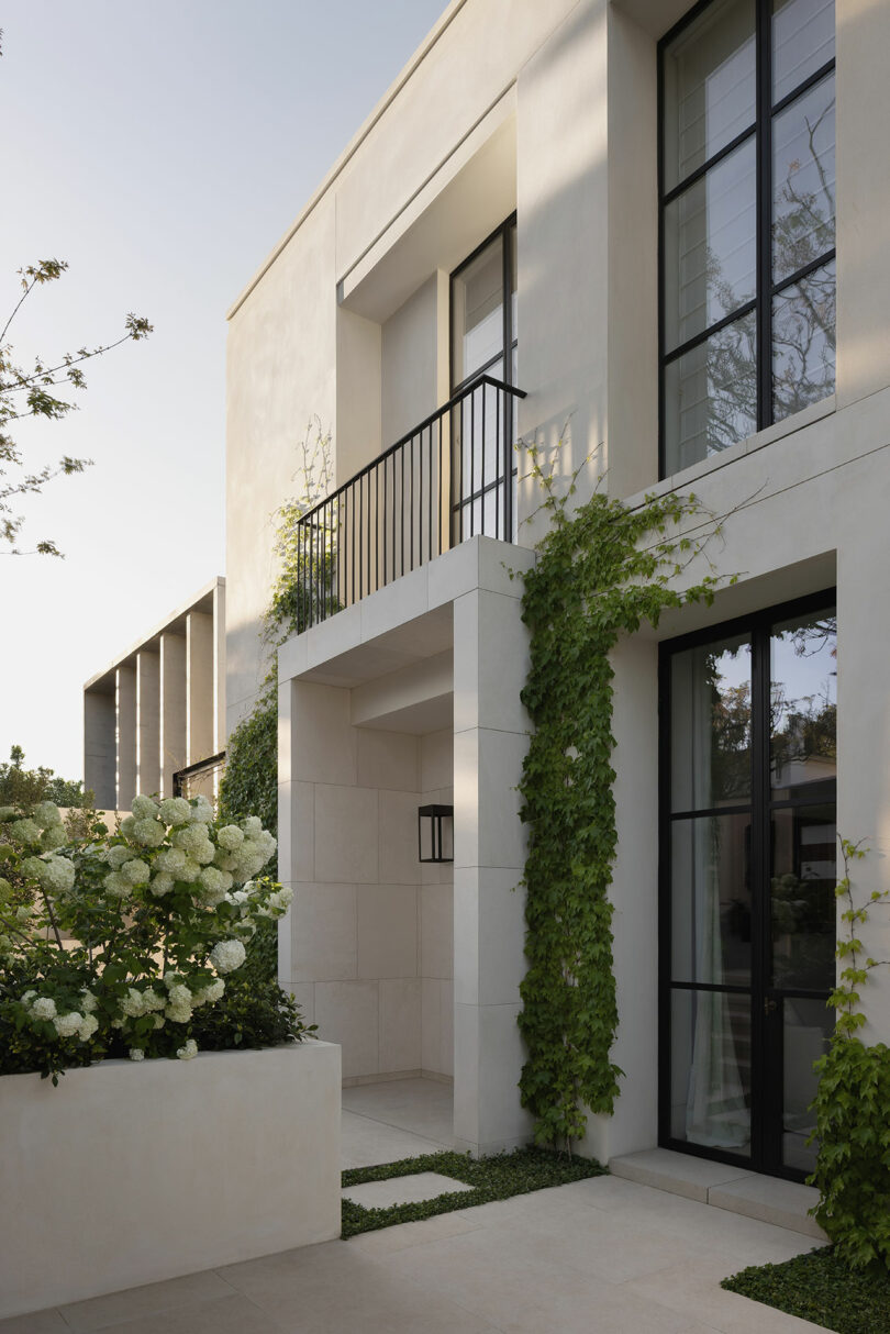 A modern, minimalist two-story house with large black-framed windows, light-colored exterior walls, and climbing greenery along the facade.