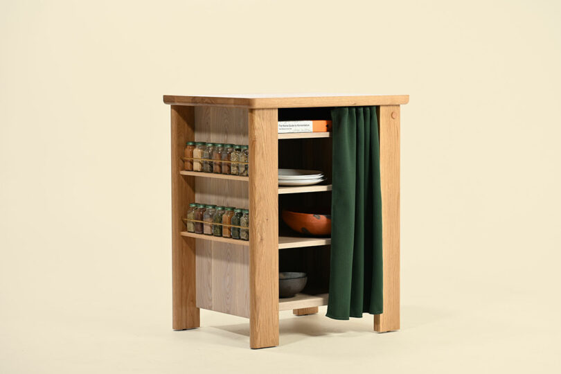 Una pequeña isla de cocina de madera con estantes abiertos que contienen platos, tazones, libros y frascos, con una cortina verde que cubre parcialmente un lado.