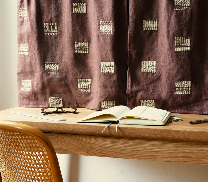 Una mesa de madera con un cuaderno abierto, un bolígrafo y unas gafas, y una cortina estampada delante de la ventana. Debajo de la mesa hay una silla tejida.