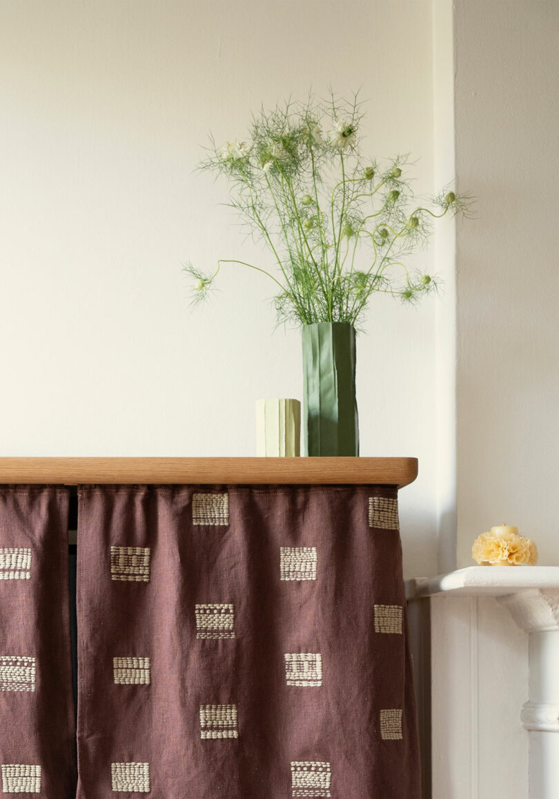 Un jarrón verde con escasa vegetación y un pequeño libro se encuentran sobre una superficie de madera cubierta con una tela marrón estampada. Una flor amarilla descansa sobre una repisa cercana.