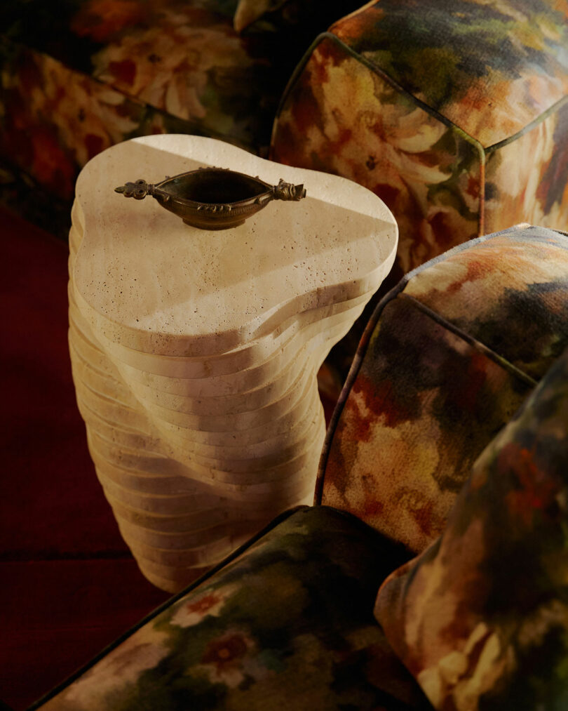 Una mesa auxiliar de piedra tallada con formas orgánicas en capas sostiene un pequeño plato decorativo, colocado entre sillas tapizadas con motivos florales.