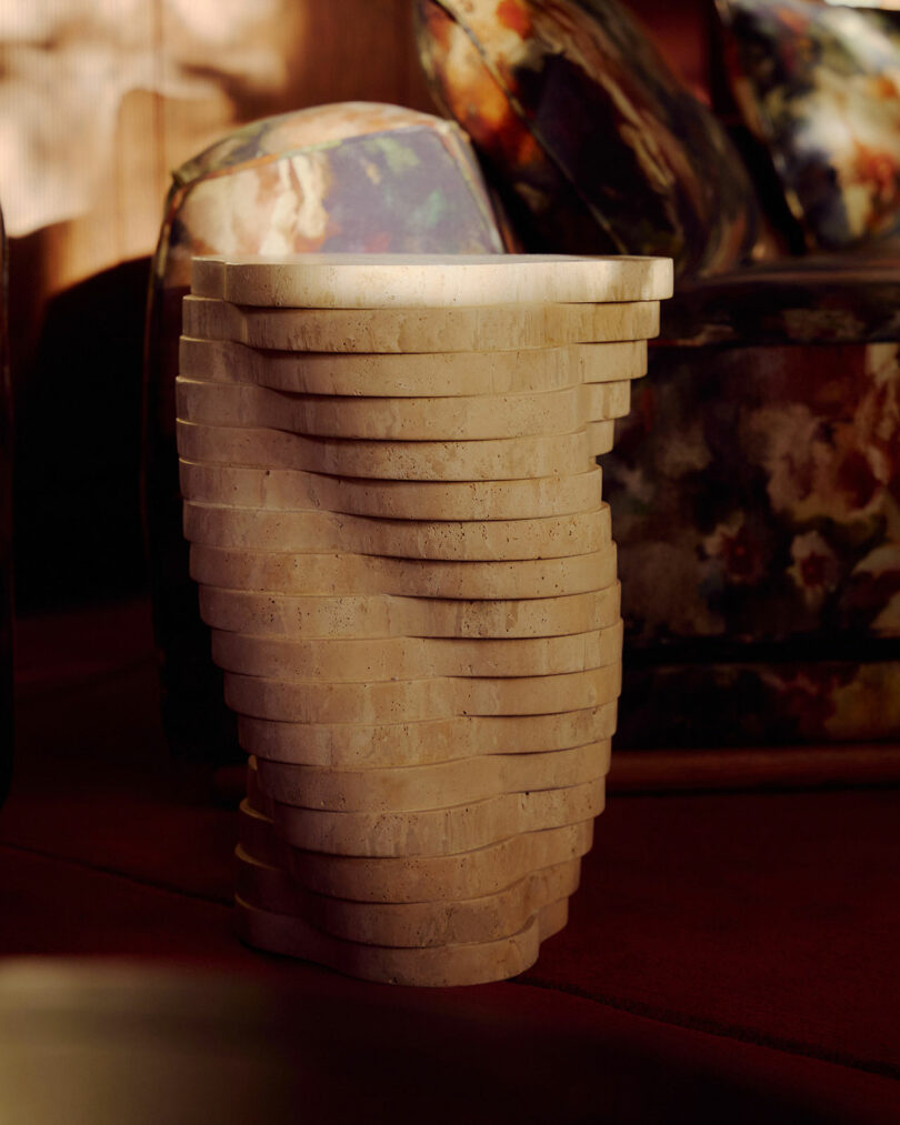 Una escultural mesa auxiliar escalonada de piedra beige con un diseño retorcido se asienta sobre un piso rojo pálido, junto a una silla con estampado floral.