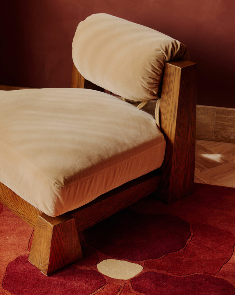 Un sillón de madera con asiento y respaldo tapizados en color beige descansa sobre una alfombra con motivos florales rojos sobre paredes de color marrón rojizo.