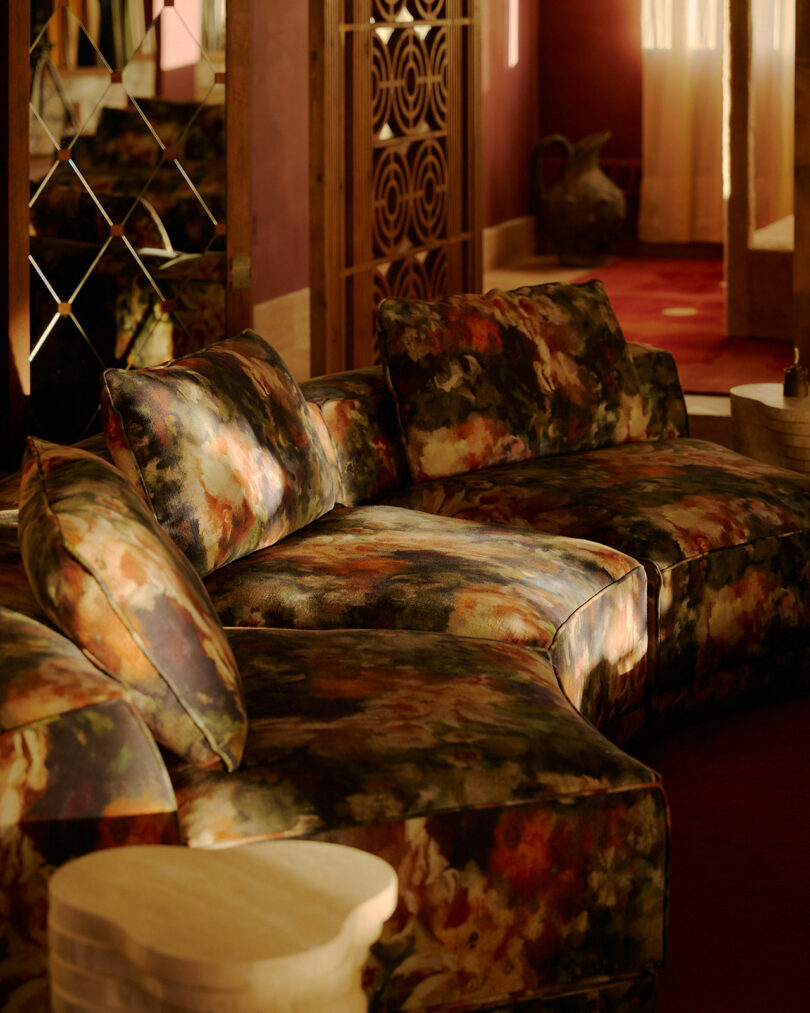 Un sofá seccional con estampado floral se encuentra en una habitación soleada con paredes rojas, paneles de espejos y detalles en madera.
