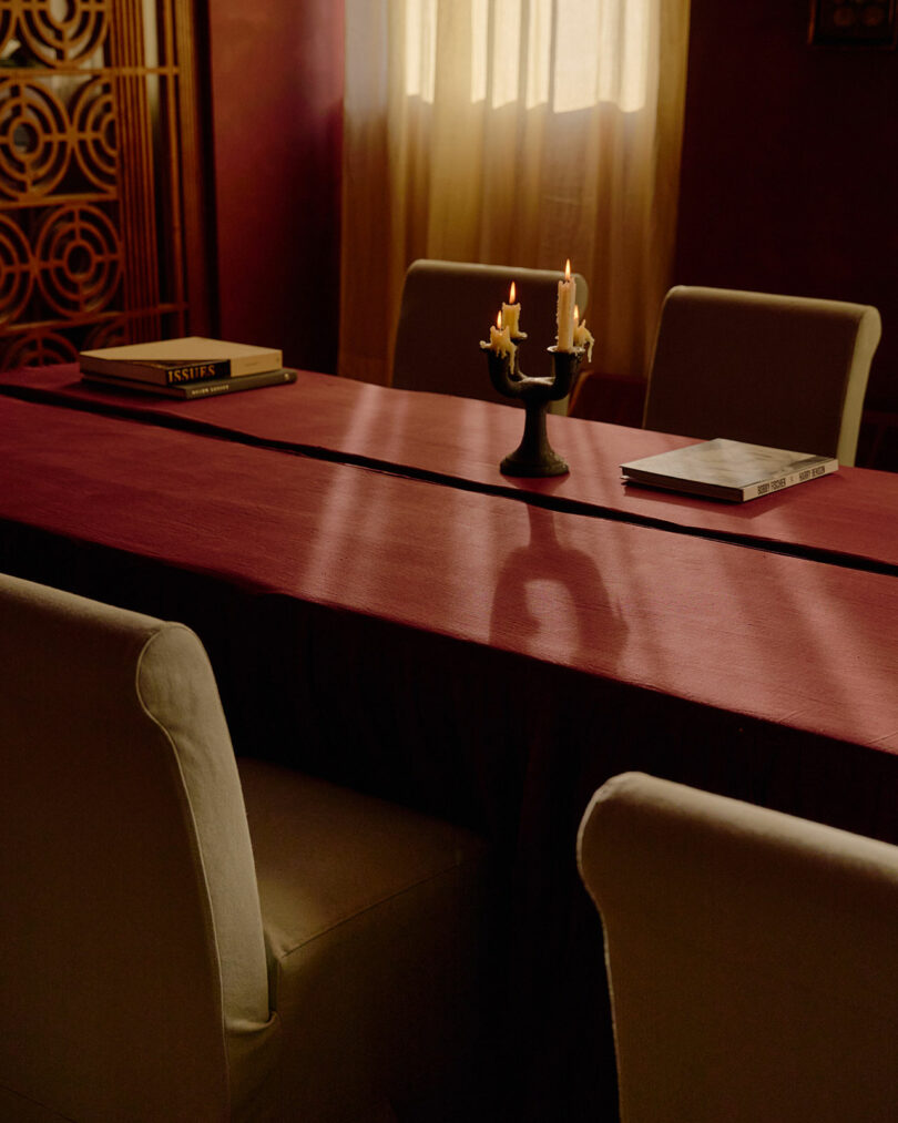 La habitación con poca luz está cubierta con un mantel granate, cuatro sillas blancas, un candelabro con velas derretidas y dos libros sobre la mesa cerca de una ventana con cortinas transparentes.