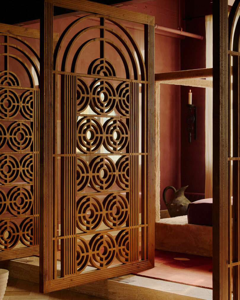 Un biombo decorativo de madera con un patrón circular geométrico se alza en un interior cálido con un telón de fondo de paredes de barro y vasijas de barro.