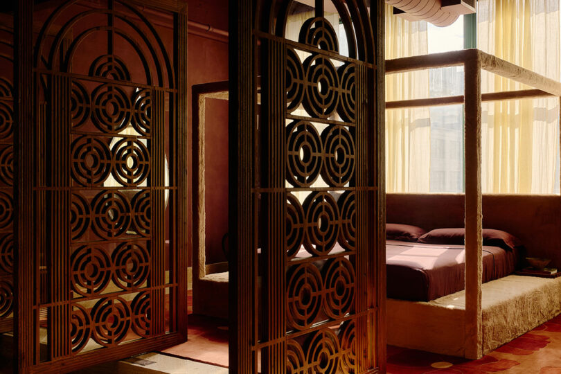 La habitación tiene biombos metálicos decorativos, una cama con dosel con ropa de cama de color violeta y cortinas amarillas que filtran la luz del sol a través de las ventanas.