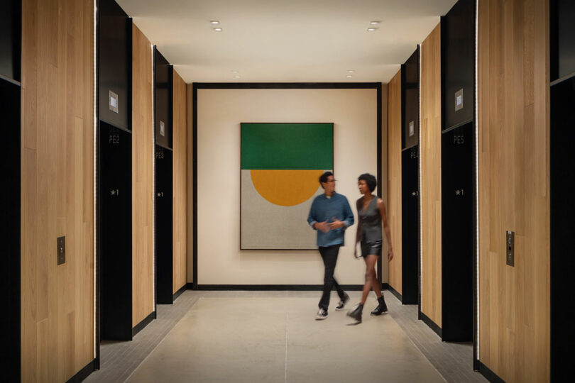 Dos personas caminan por un pasillo moderno bordeado de puertas de ascensor y que termina con una obra de arte geométrica: un espacio interior diseñado por Maurice Adjemi Architects.