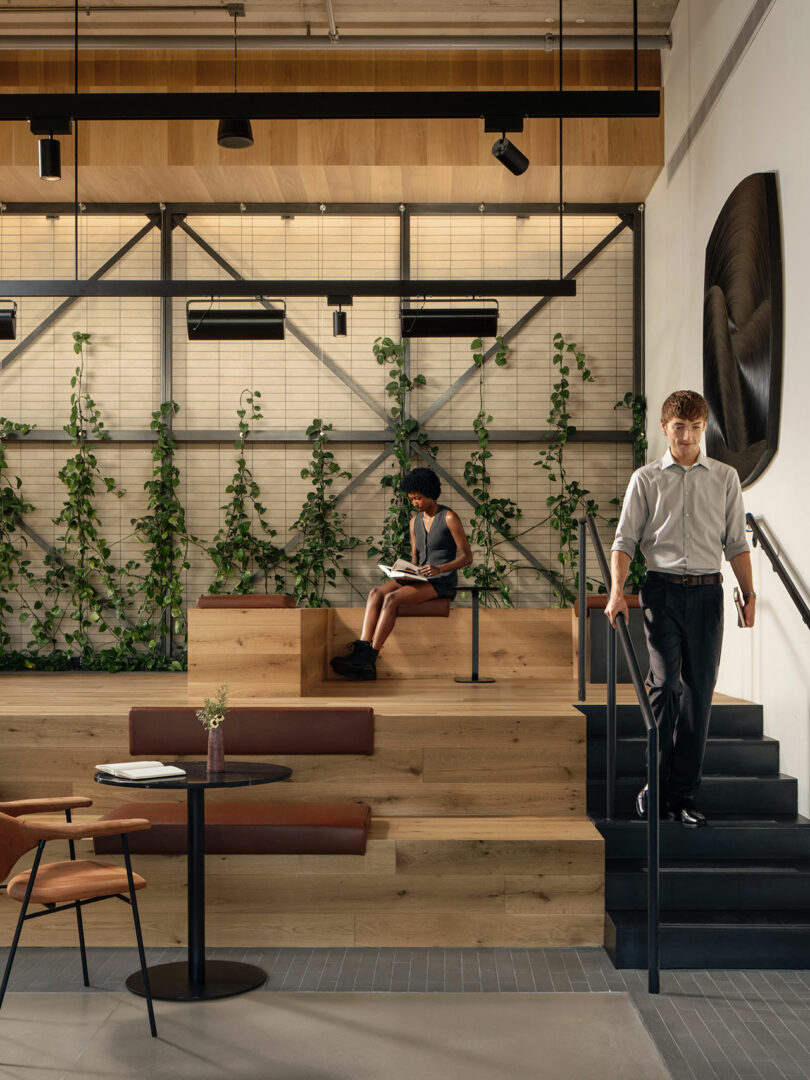 Morris Adjmi Architects diseñó un interior moderno lleno de plantas donde un hombre baja las escaleras mientras otro se sienta en un asiento de madera escalonado leyendo un libro.