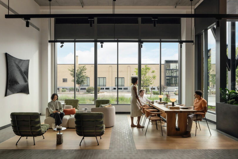 Diseñado por Morris Adjmi Architects, el moderno salón de oficina cuenta con grandes ventanales donde cuatro personas interactúan en sofás y sillas, mientras que una larga mesa de madera y exuberantes plantas realzan la belleza del espacio.