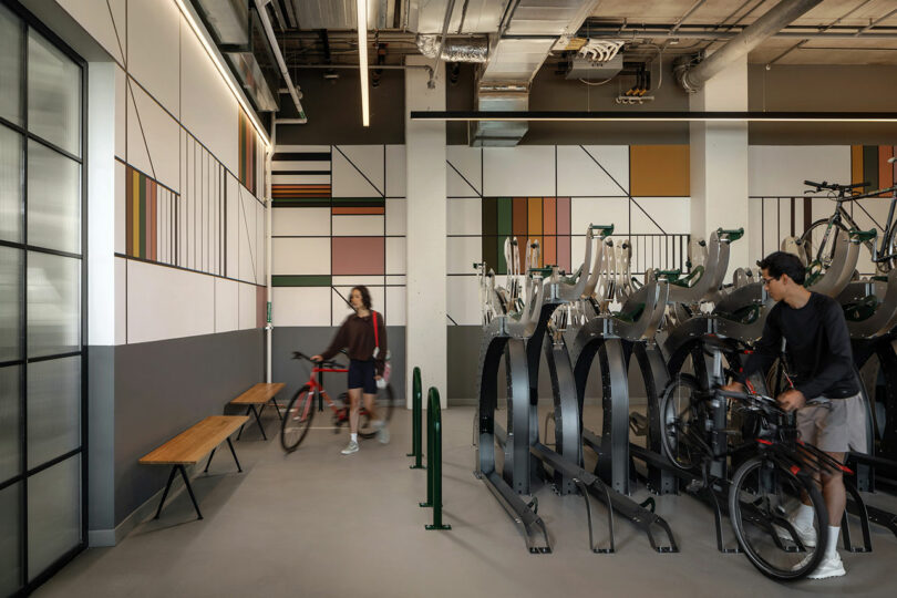 Dos ciclistas se encuentran en una moderna sala interior para guardar bicicletas con arte geométrico en las paredes, bancos y portabicicletas diseñados por Morris Adjmi Architects.