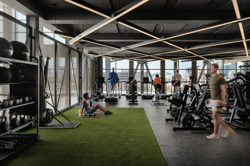El moderno gimnasio diseñado por Morris Adjmi Architects cuenta con grandes ventanales, varias personas usando equipos cardiovasculares y de fuerza, y una persona sentada en césped artificial junto al equipo de ejercicio.