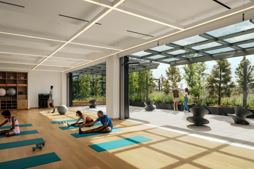 El moderno gimnasio diseñado por Morris Adjmi Architects cuenta con grandes ventanales, con personas estirándose sobre colchonetas de yoga en el interior y dos en la terraza al aire libre con vegetación y asientos esculturales. La luz natural llena todo el espacio.