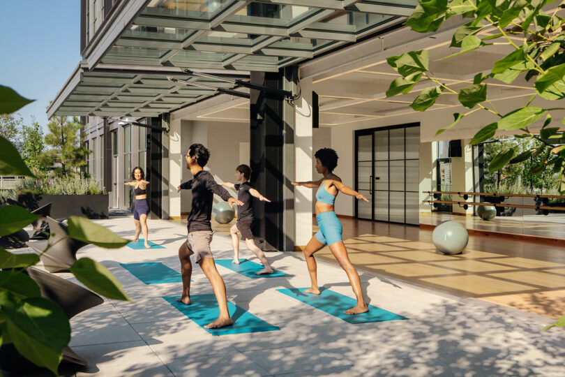 Cuatro personas practican yoga sobre colchonetas azules frente a un edificio moderno con grandes puertas de vidrio diseñado por Maurice Adjemi Architects. Una pelota de ejercicio cubierta y la vegetación circundante contribuyen a la atmósfera tranquila.