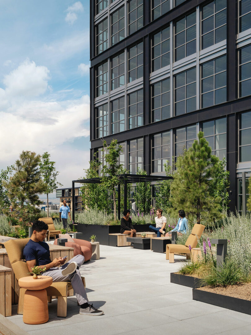 La gente se relaja con modernos muebles de exterior en la terraza de la azotea, rodeada de vegetación, junto a un alto edificio con paneles de vidrio diseñado por Maurice Adjemi Architects.