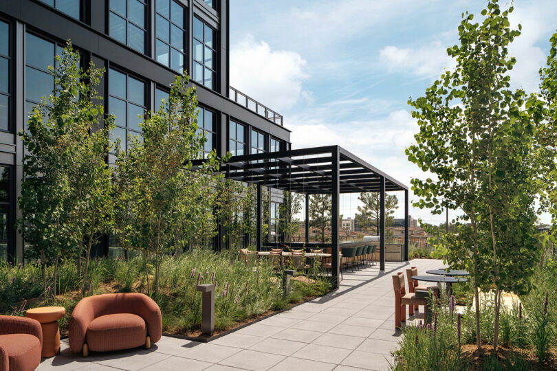 Una moderna terraza al aire libre diseñada por Morris Adjmi Architects cuenta con áreas para sentarse, mesas, vegetación y una pérgola junto a un edificio de oficinas de vidrio bajo un cielo parcialmente nublado.