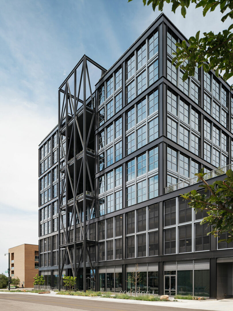 Moderno edificio de oficinas de varias plantas diseñado por Morris Adjmi Architects con fachadas de acero y vidrio negro, soportes estructurales exteriores y grandes ventanales rodeados de varios árboles bajo un cielo despejado.
