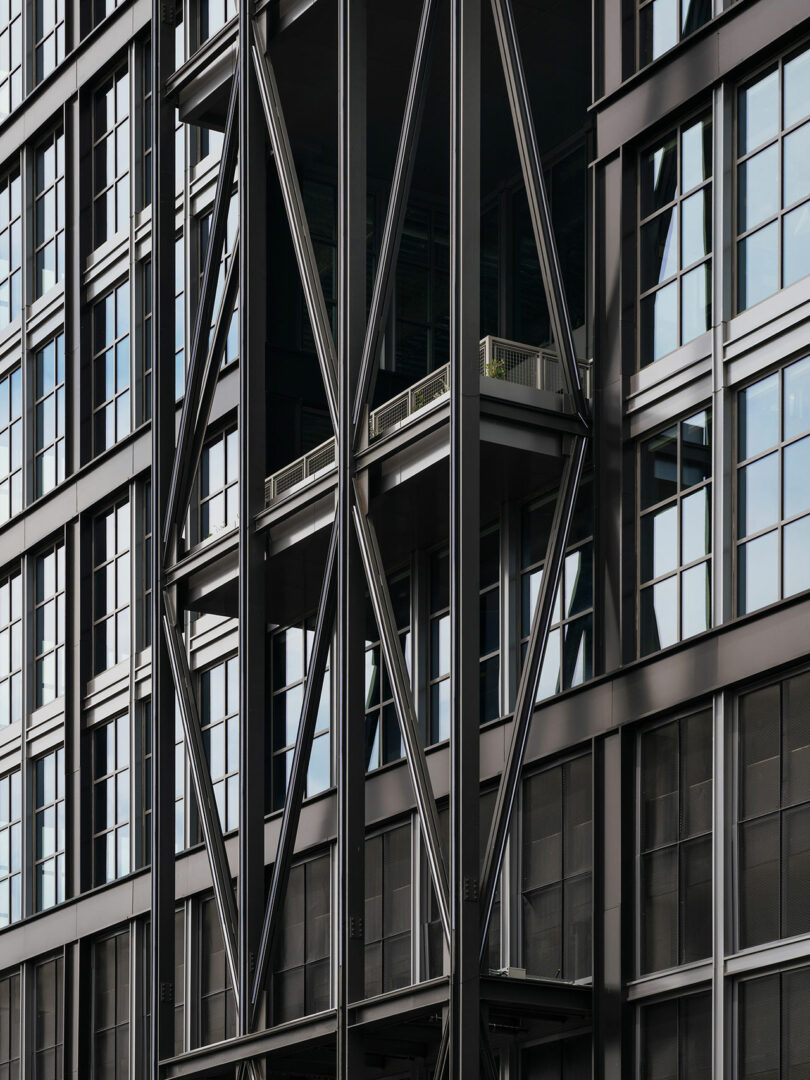 La moderna fachada del edificio diseñada por Morris Adjmi Architects presenta ventanas de vidrio, vigas de metal negro en forma de X y balcones con pequeñas barandillas y vegetación.