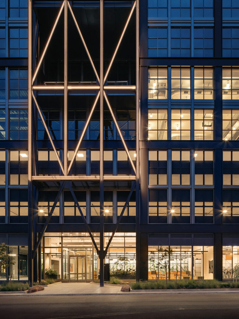Fachada de edificio moderno al atardecer con luces lineales que forman patrones geométricos que reflejan el estilo característico de Maurice Adjemi Architects; Las ventanas de vidrio revelan los luminosos espacios interiores y la entrada principal al nivel de la calle.