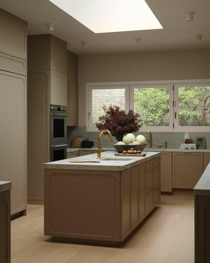 Modern kitchen with beige cabinets, marble countertops, and a central island featuring a gold faucet—reflecting OSKLO’s signature elegance. Large windows overlook greenery, while a vase of flowers and a bowl of onions adorn the island.
