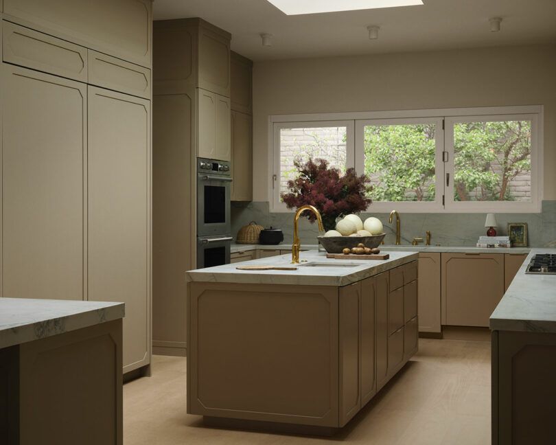 Modern OSKLO kitchen with beige cabinets, marble countertops, a central island, gold fixtures, built-in ovens, and a large window overlooking greenery.