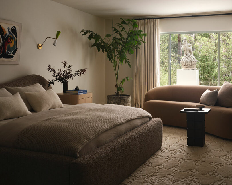 A modern OSKLO bedroom with a large bed, sofa, potted plants, abstract wall art, and a window revealing an outdoor statue surrounded by greenery.
