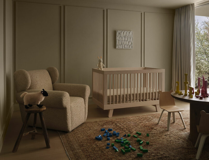 A neutral-toned OSKLO nursery with a crib, plush chair, wooden stool with a toy sheep, rug, scattered building blocks, table, chairs, and toys arranged near a curtained window.