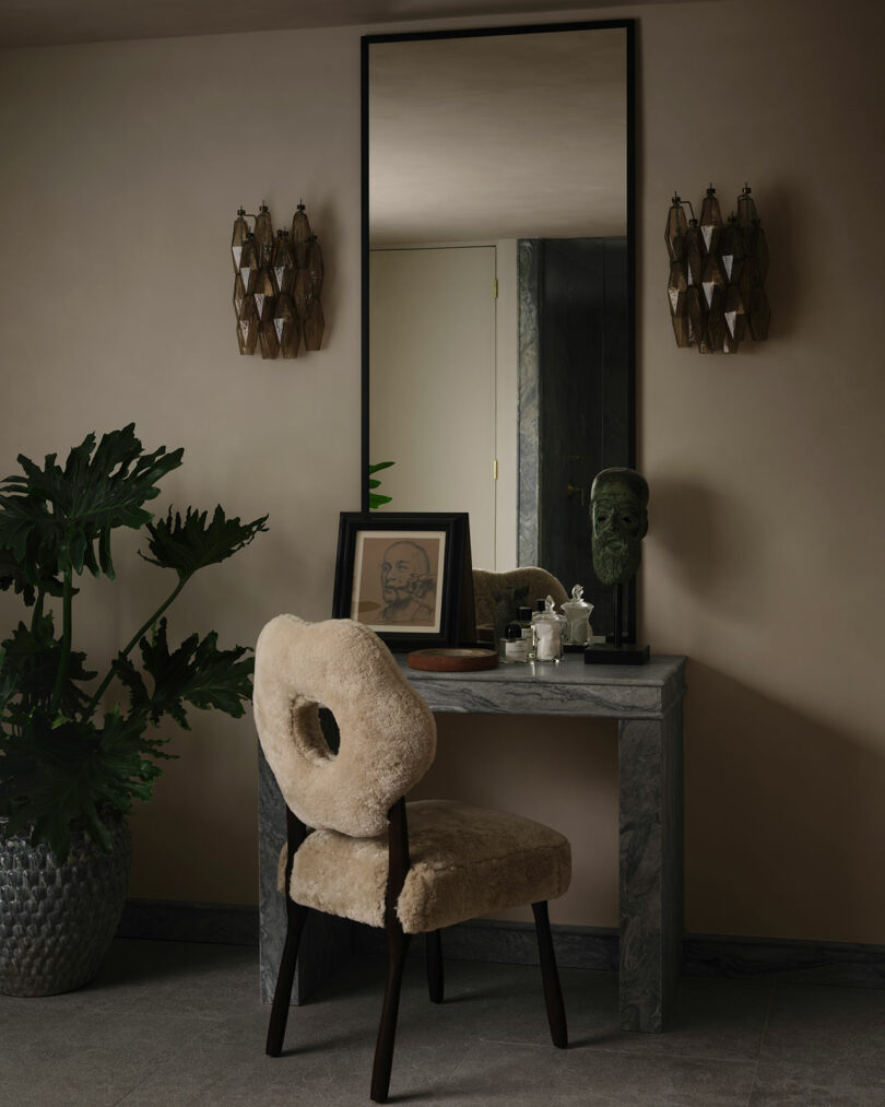 An OSKLO vanity with a marble top, large framed mirror, plush chair, framed artwork, decorative items, and wall sconces sits against a neutral-toned wall with a potted plant nearby.