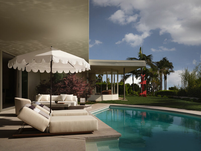 Modern OSKLO outdoor patio with white lounge chairs and umbrella beside a curved swimming pool, surrounded by garden, palm trees, and a covered seating area in the background under a partly cloudy sky.