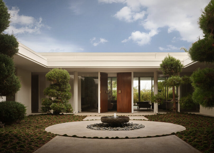 Modern courtyard by OSKLO with circular landscaping, a central stone fountain, and a minimalist building entrance featuring large wooden doors and glass walls under a partly cloudy sky.