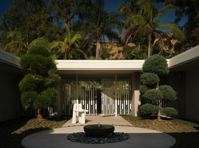 A modern OSKLO courtyard with sculpted trees, a circular stone fountain, a white abstract chair, and a gate in the background, all surrounded by palm trees.