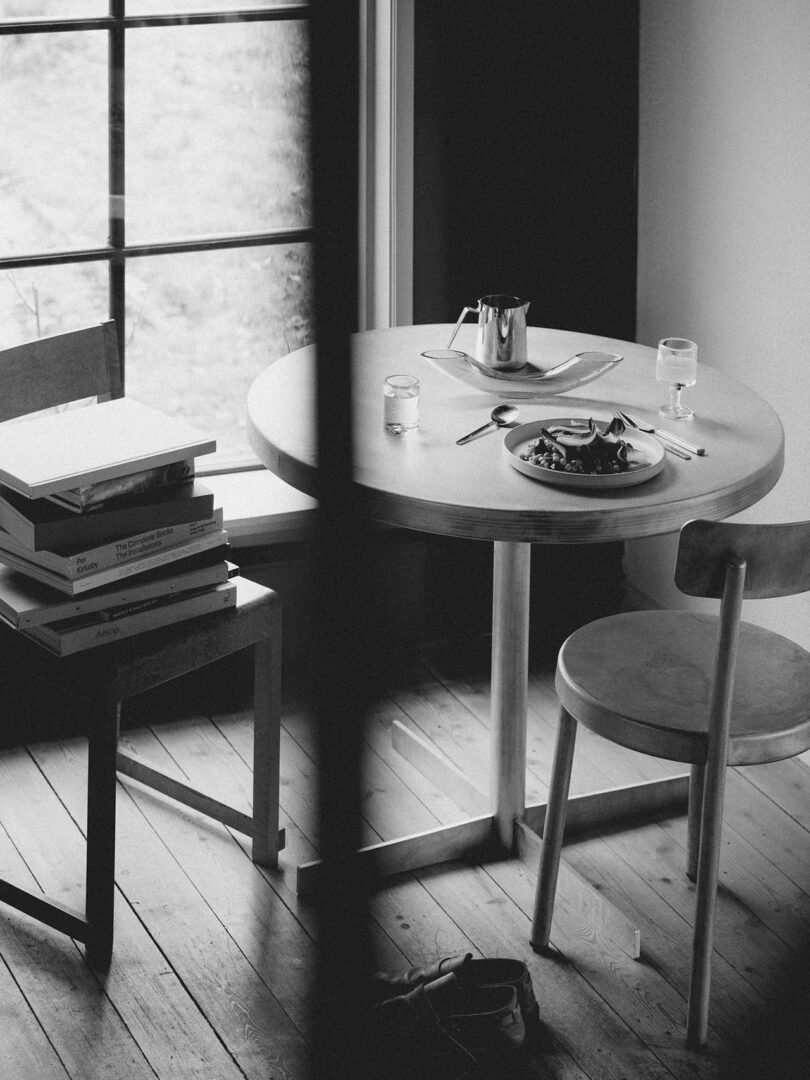 Una mesa de TASCA FRAMA está rodeada por un plato de comida, cubiertos, un vaso de agua y una tetera, junto a dos sillas, una de las cuales está cerca de una pila de libros junto a la ventana. Foto en blanco y negro.