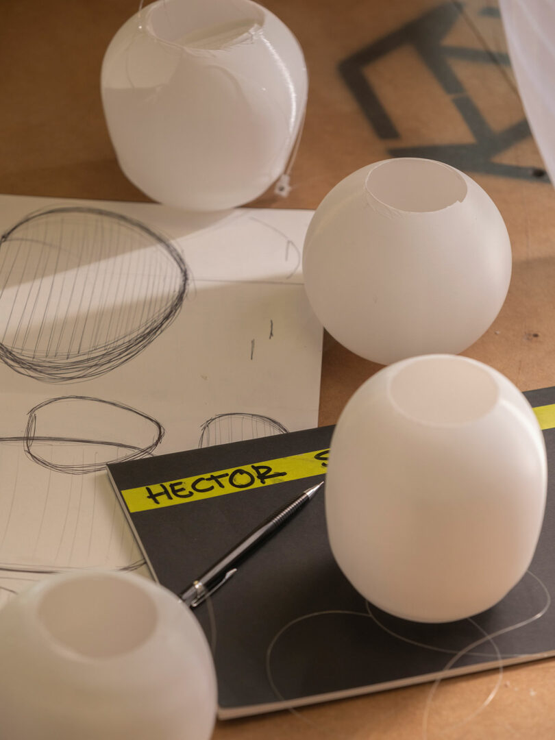 Four white spherical objects are placed on a table with sketchpads showing sphere drawings, a pen, and a folder labeled "HECTOR" in yellow.