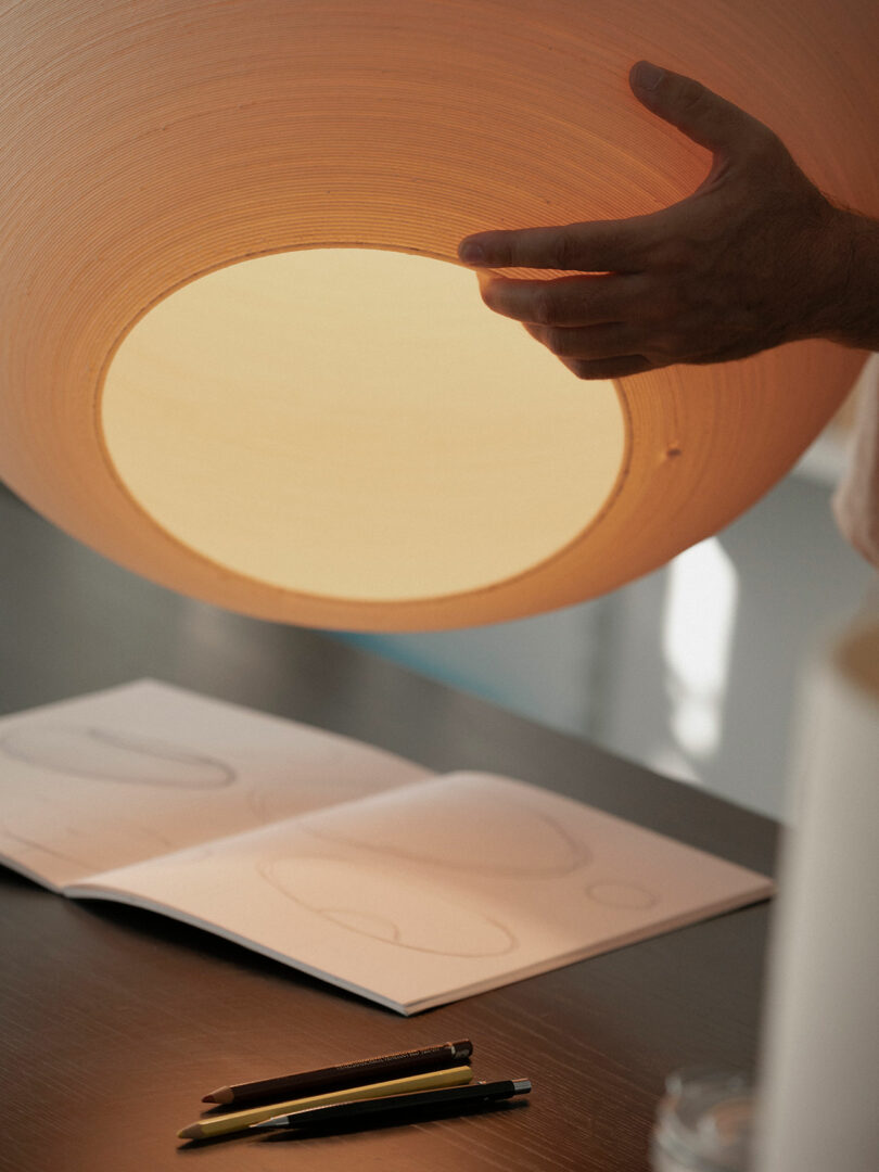 A hand adjusts a large round lampshade above a table with an open sketchbook and three pens.