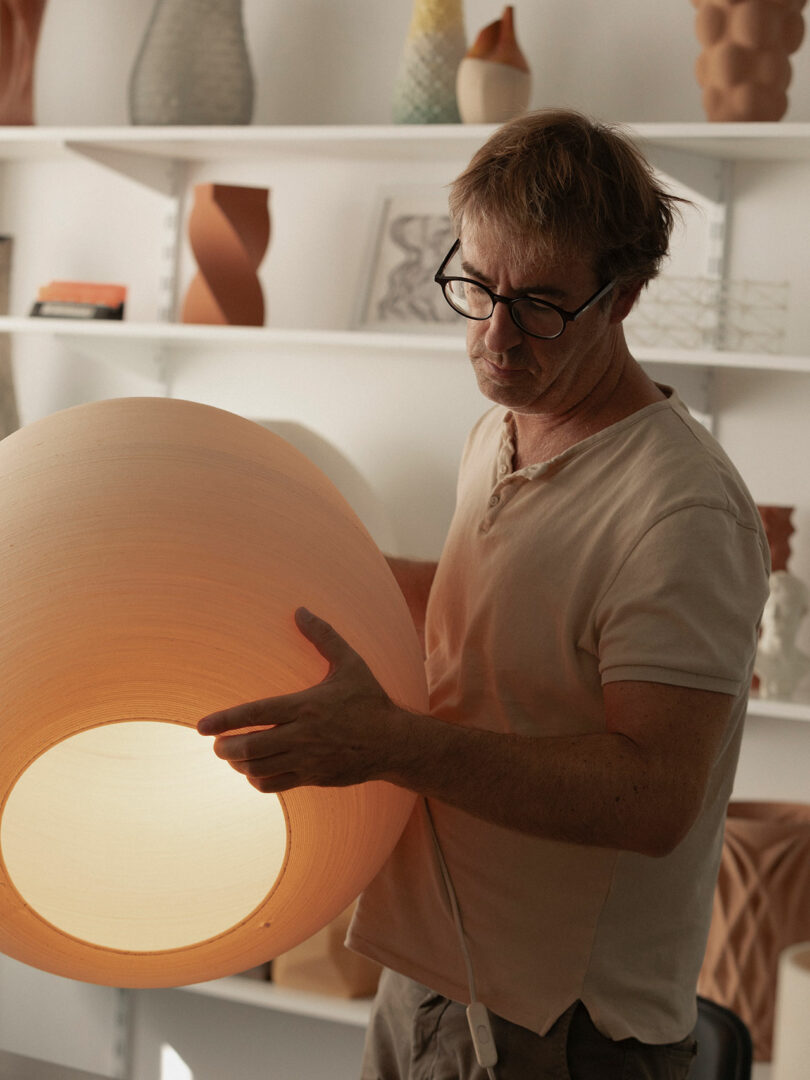 A man wearing glasses inspects a large, round, light-colored lampshade in a studio with shelves holding pottery and art objects in the background.