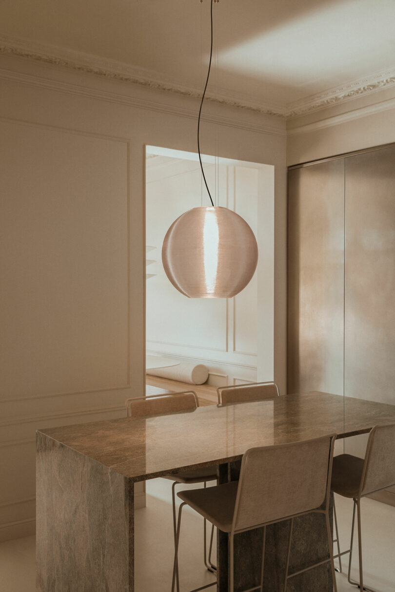 A minimalist dining area with a stone-top table, four beige chairs, and a large pendant light hanging from the ceiling. Neutral tones and soft lighting create a calm atmosphere.