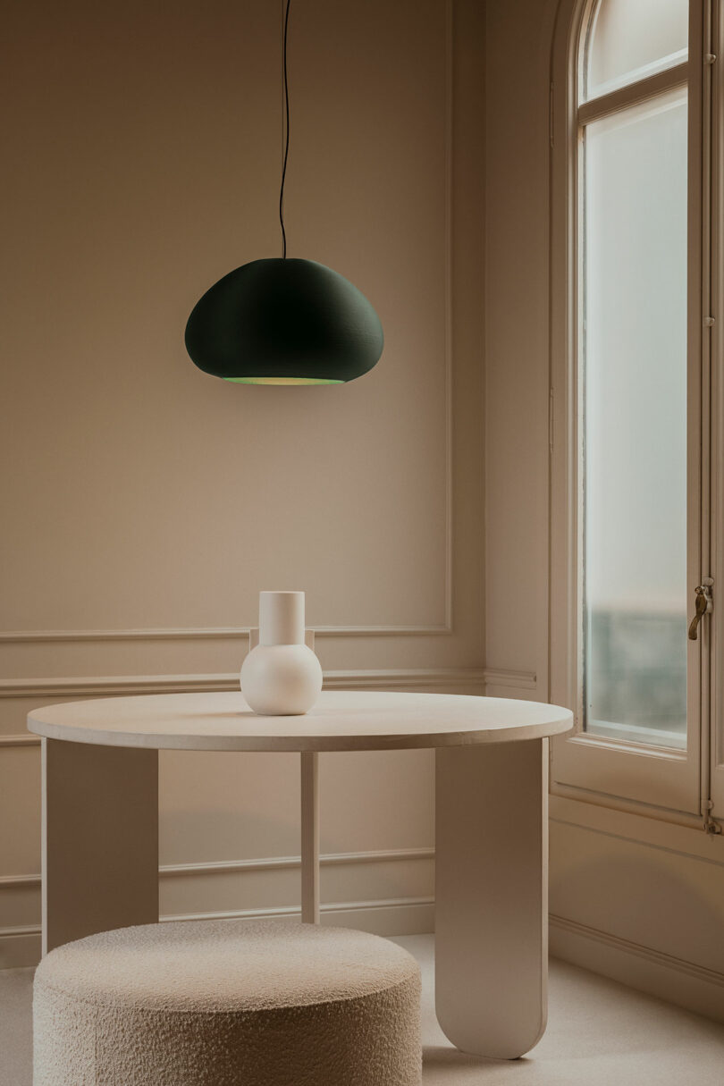 A minimalist room with a round white table, a white vase, a textured ottoman, a black pendant lamp, and a tall window letting in soft light.